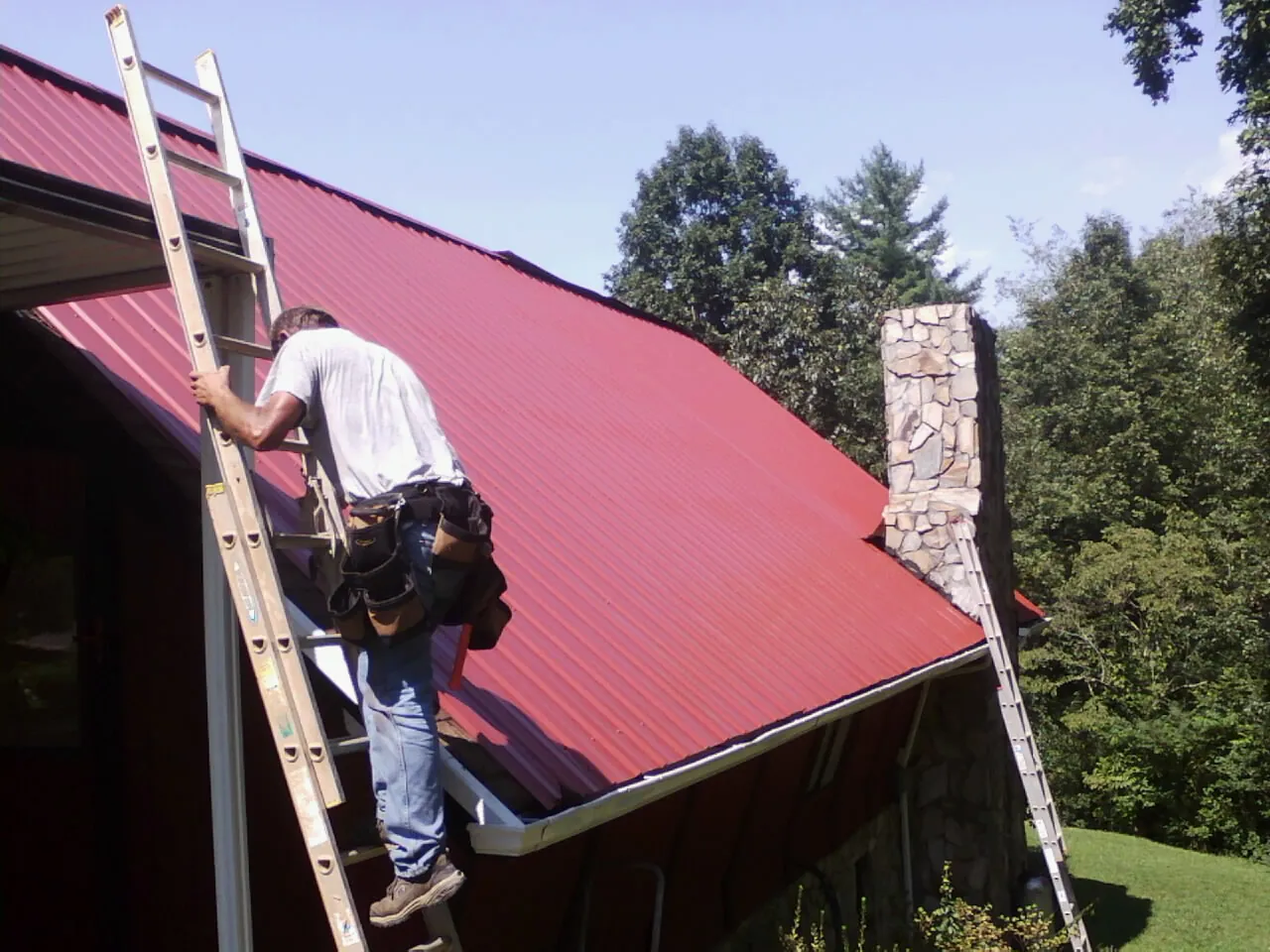 Roofer on a ladder beside a red metal roof and stone chimney — Soffit & Fascia Repair by Roofing Brothers in Roans Prairie